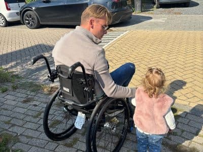 Davey reikt vanuit zijn rolstoel naar zijn dochtertje op een parkeerplaats.